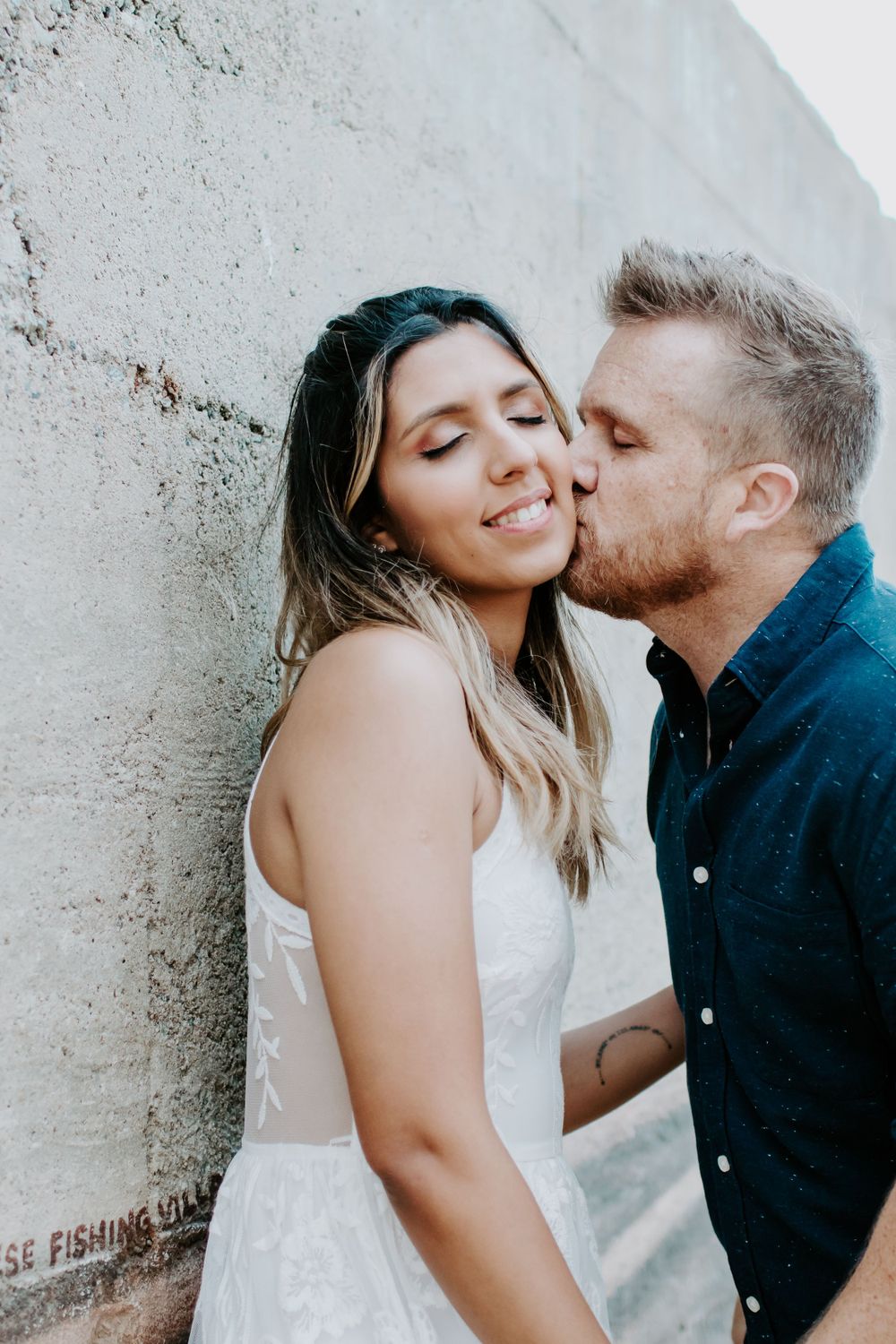 A sweet romantic moment between a couple against a white wall, with the woman in a white dress and man in navy blue.