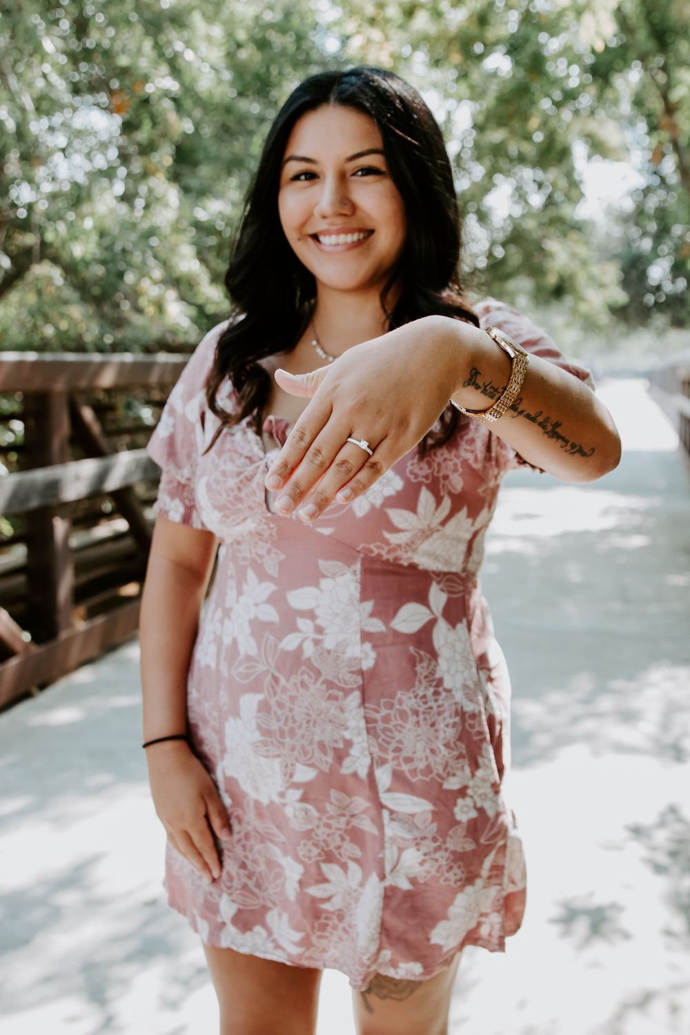 Woman in floral pink dress shows off engagement ring in sunny outdoor setting.