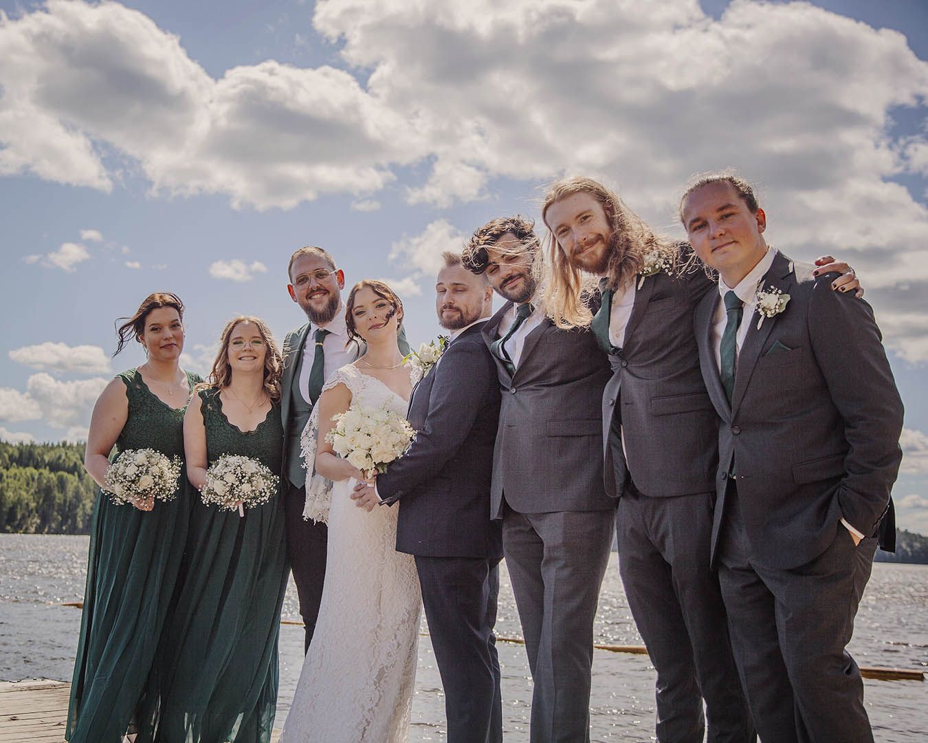 Brudfölje på brygga vid Lida friluftsområde, Stockholm. Tärnor i mörkt grönt med vita blommor, marskalkar i grå kostym.