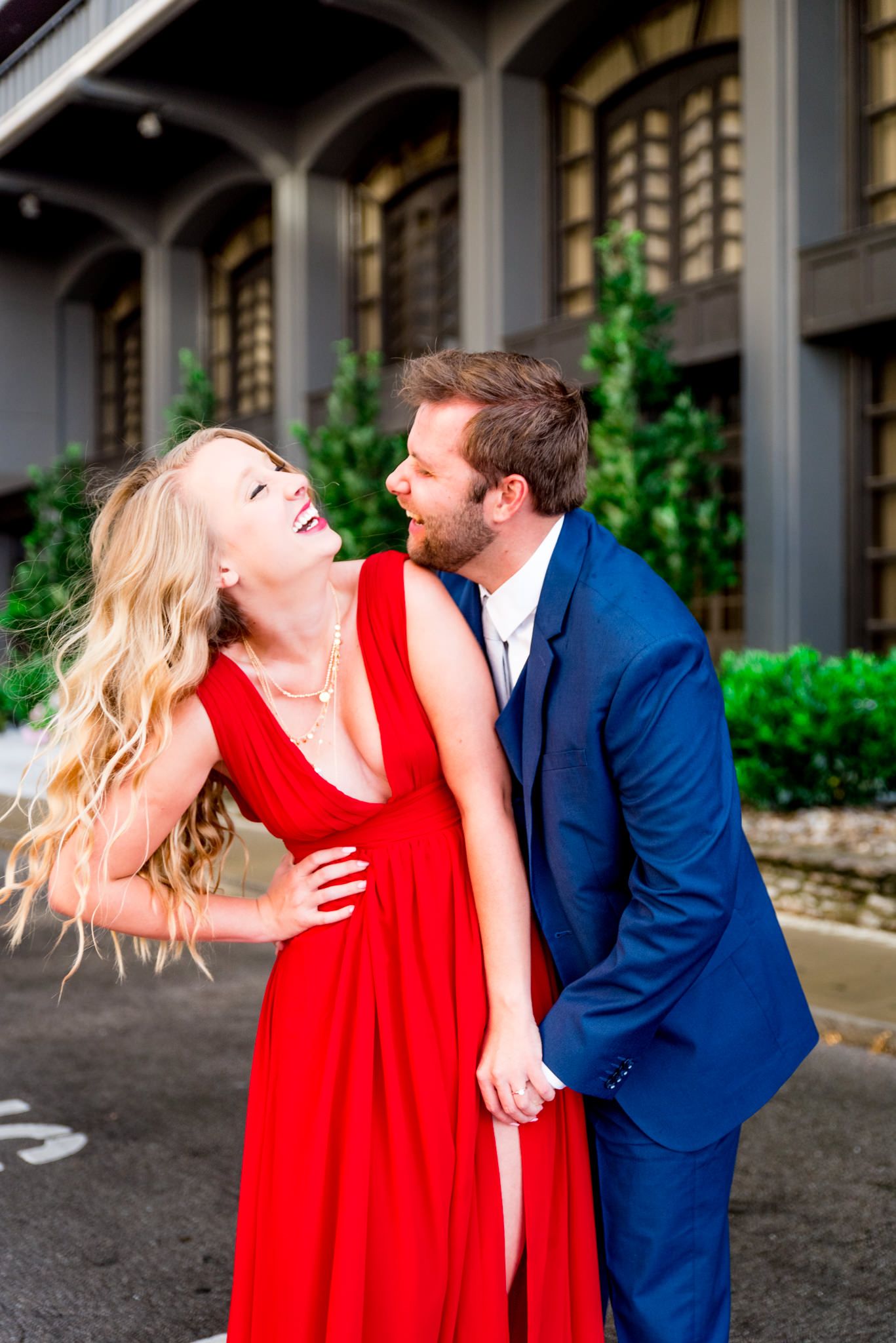 blonde woman in red dress laughing with fiance in navy blue suit tries to kiss her