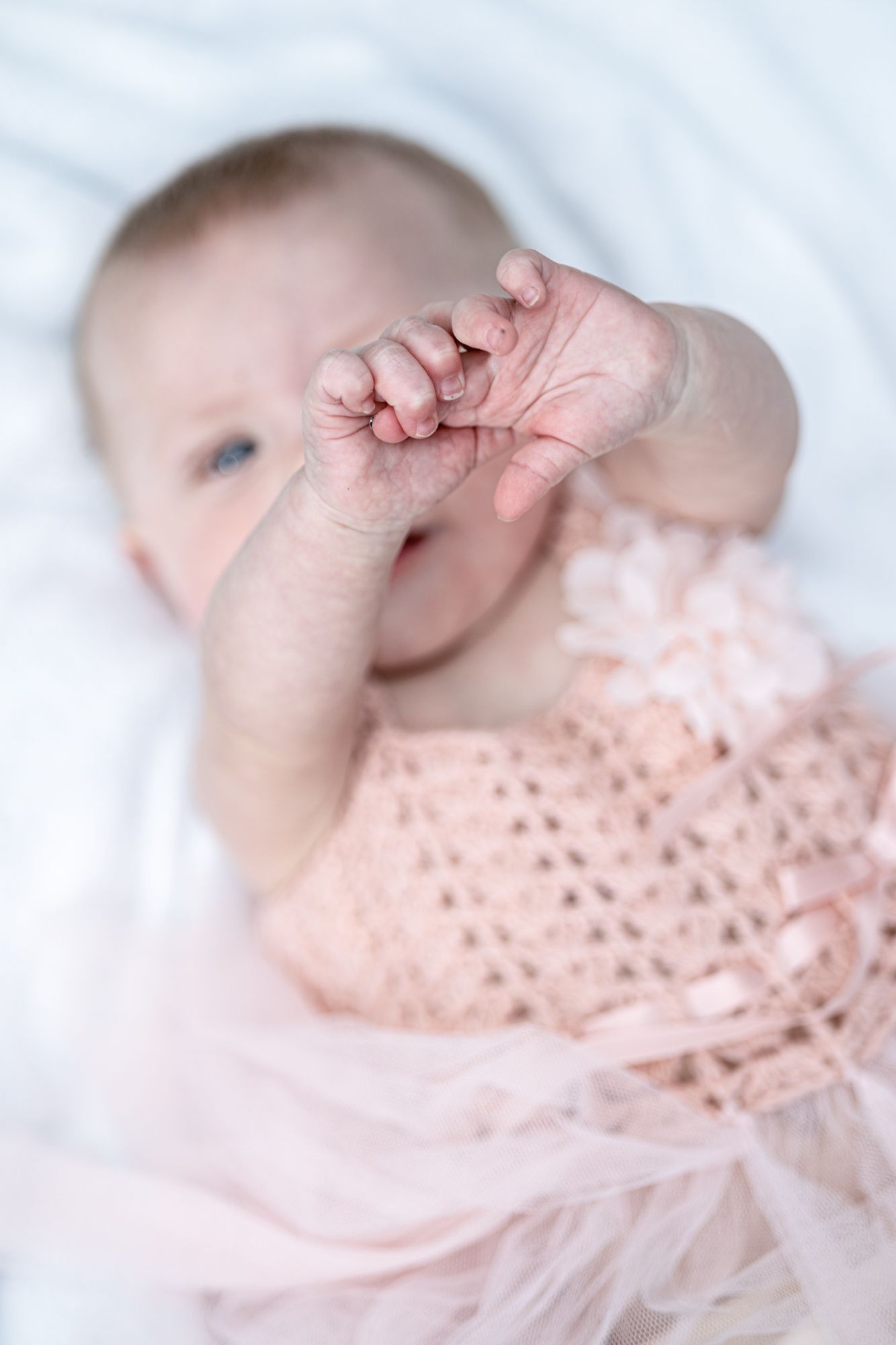 A tiny baby wearing a soft pink lace dress reaching up with their small hand against a white background.