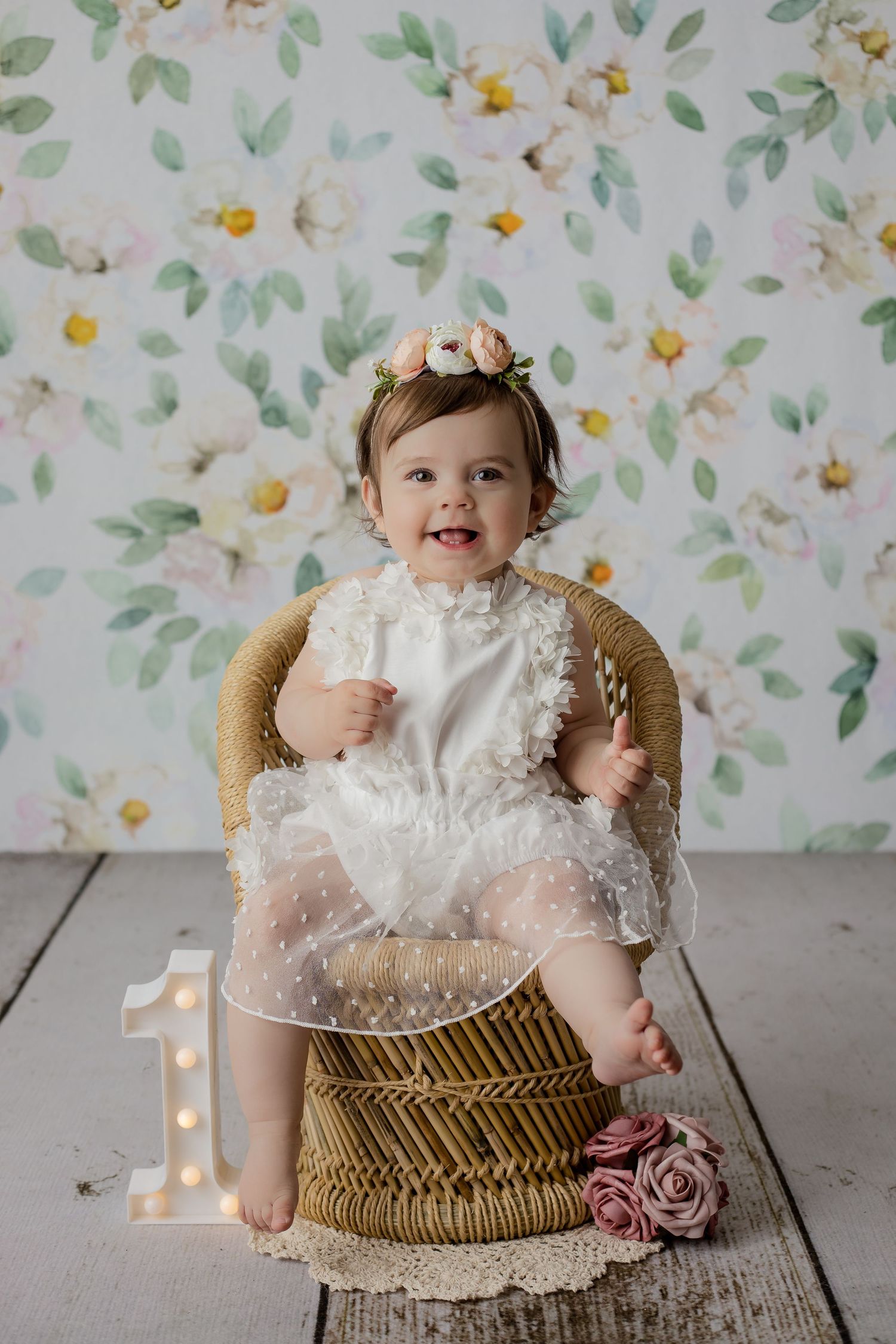 Smiling baby girl on a whicker chair and floral background in a studio setup for her first birthday photoshoot.