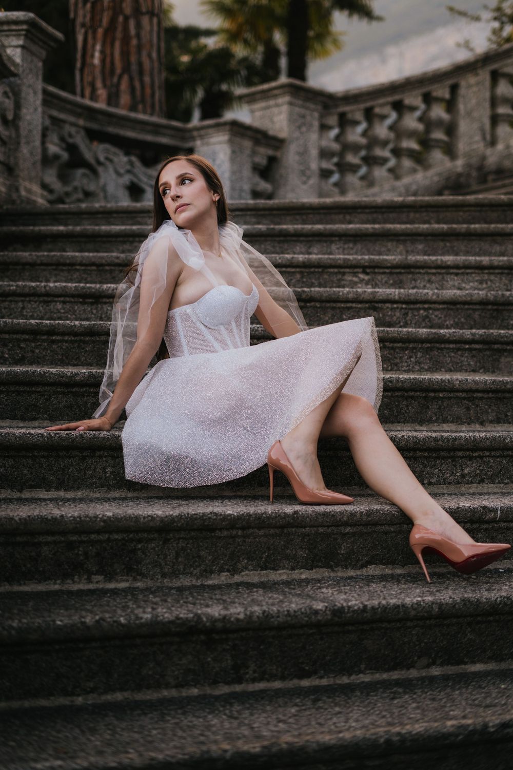 A bride in a white cocktail dress poses elegantly on stone steps with ornate railings.
