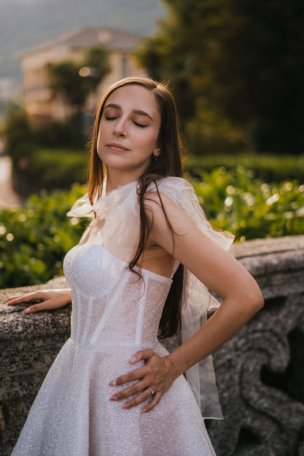 Woman in white dress poses gracefully against stone balustrade in garden setting.