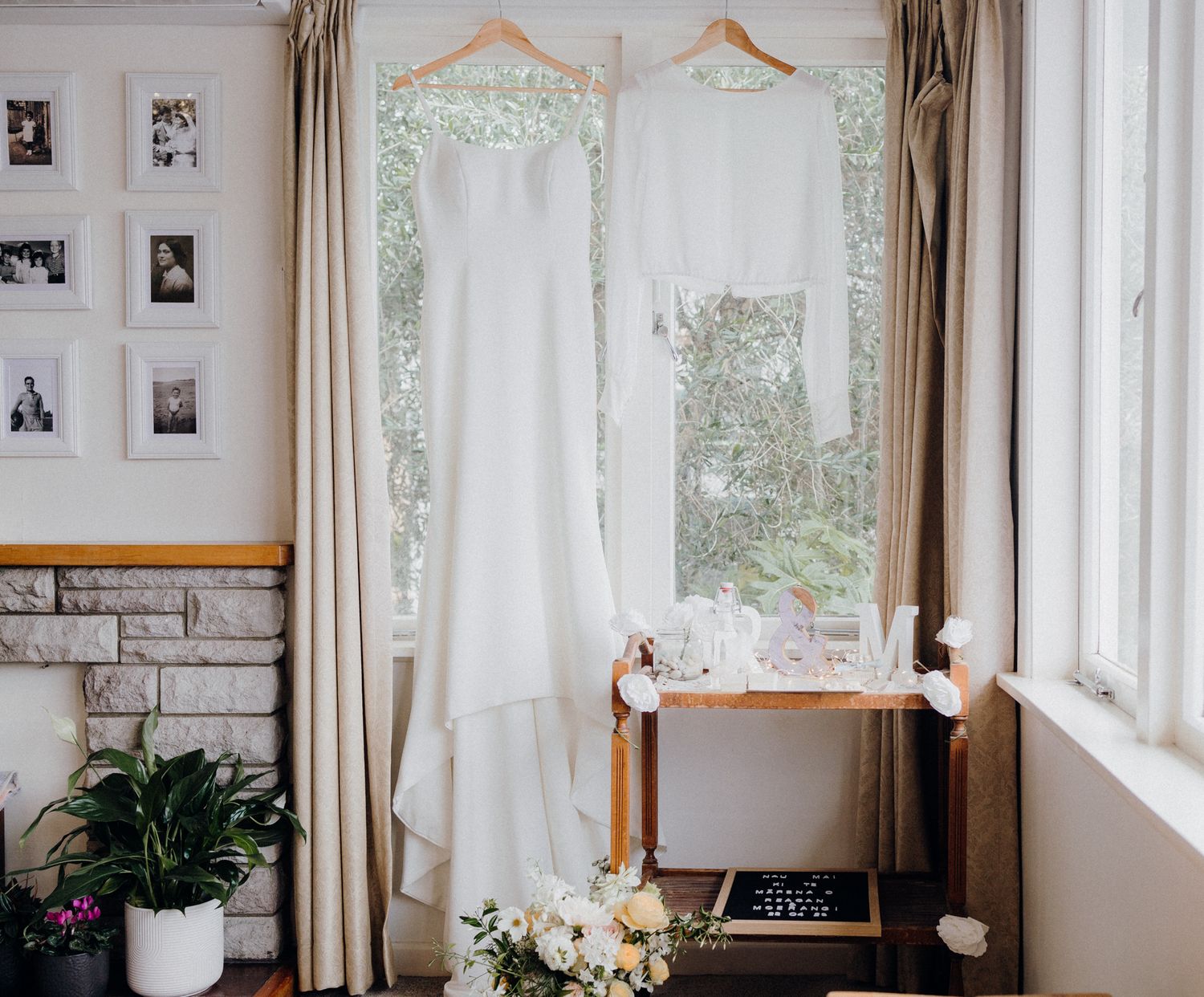 Two white wedding dresses hanging by a window with black and white photos on the wall.
