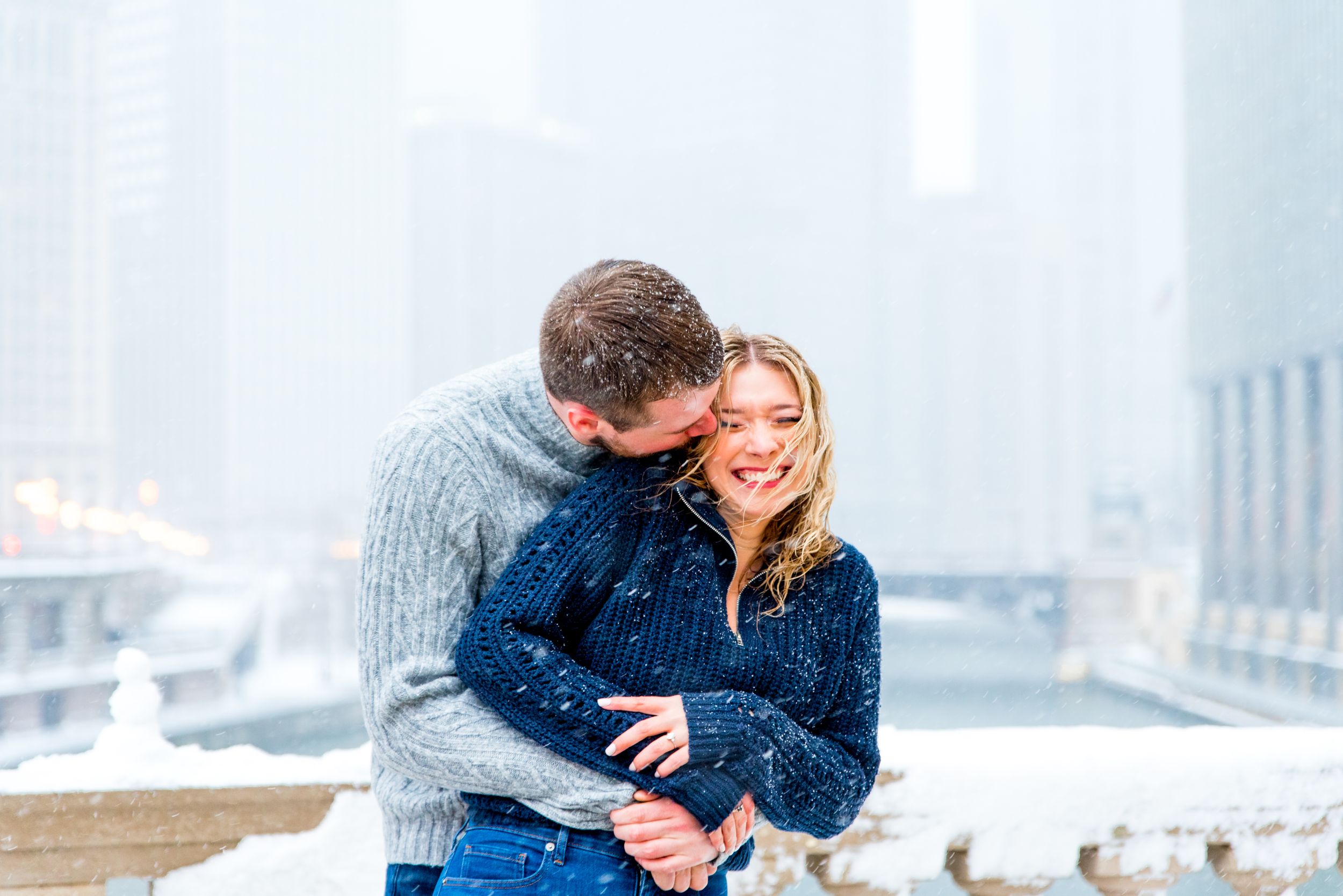 man in gray sweater hugging and kissing fiance in navy sweater at Wrigley Building for Chicago winter engagement picture