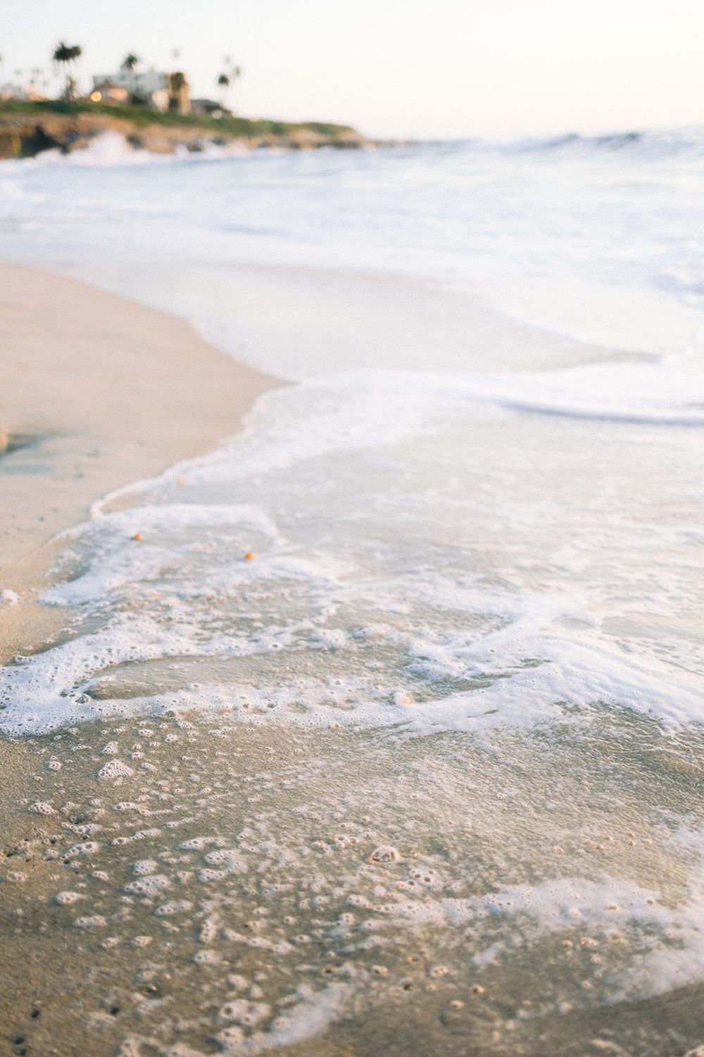 Gentle waves washing up on sandy beach shore during sunset with rocky coastline in background.