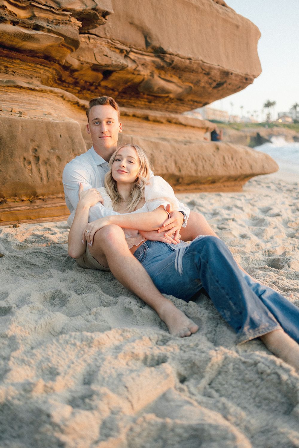 Couple relaxing on sandy beach against dramatic sandstone cliffs during golden hour.