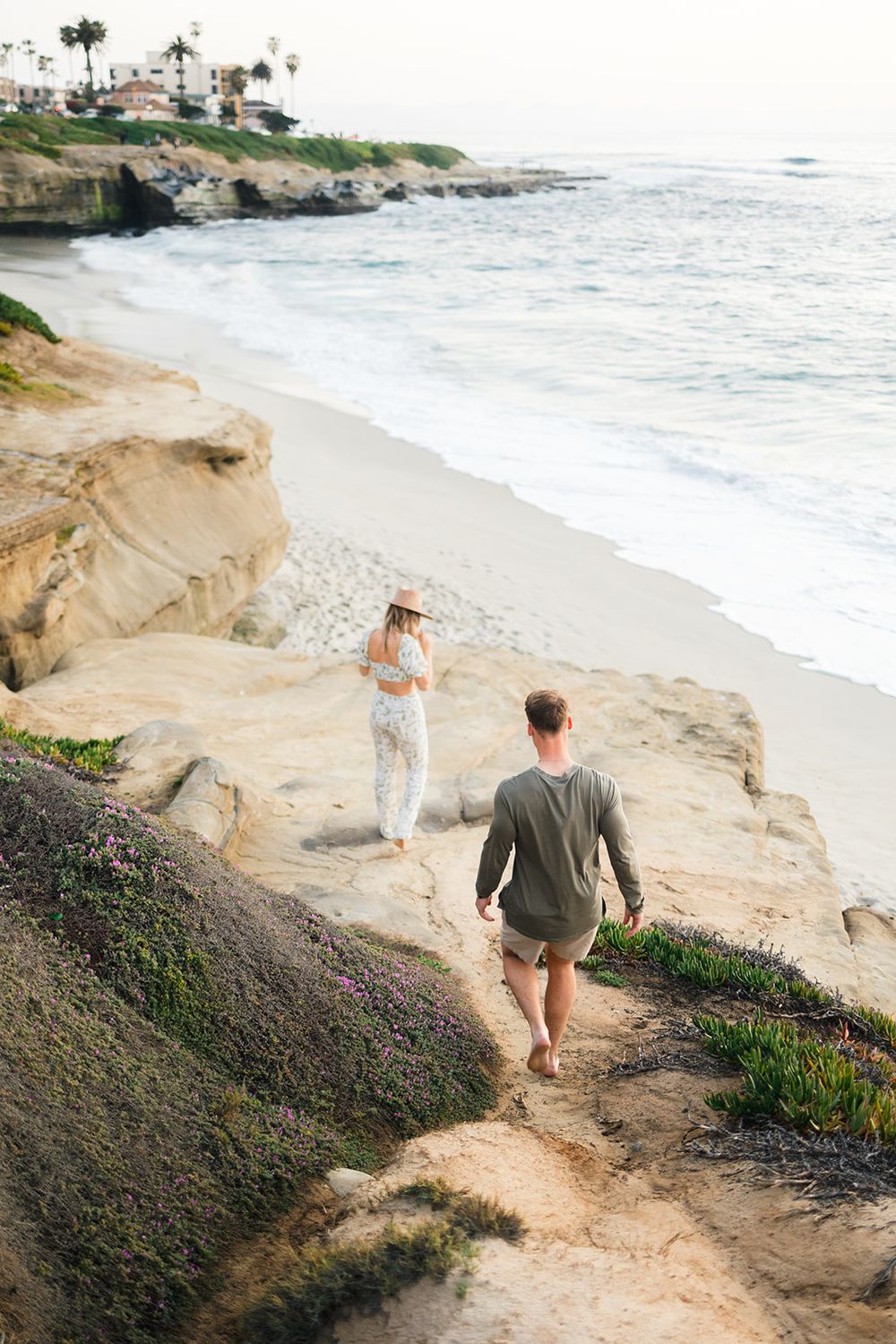 Couple walking along scenic coastal cliffs at sunset with ocean waves crashing on beach below.