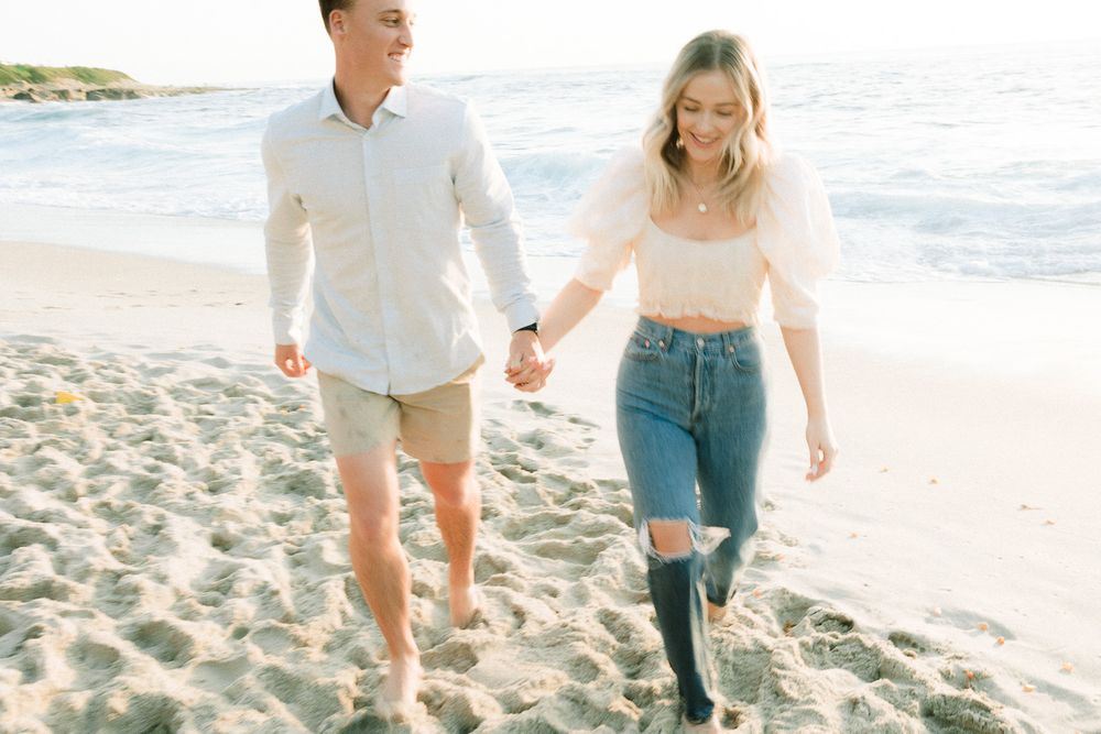 A couple walks hand in hand on a sunny beach with waves crashing in the background.