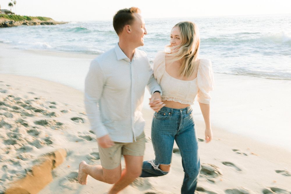 A romantic beach proposal scene with one person kneeling in the sand at sunset.