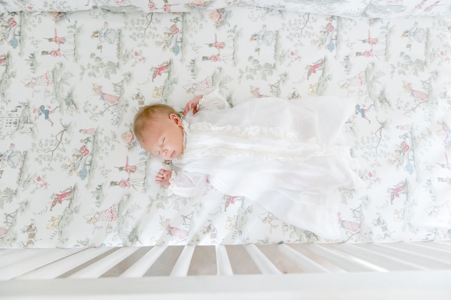 Newborn baby asleep in his crib with Peter Rabbit sheets in his christening gown.