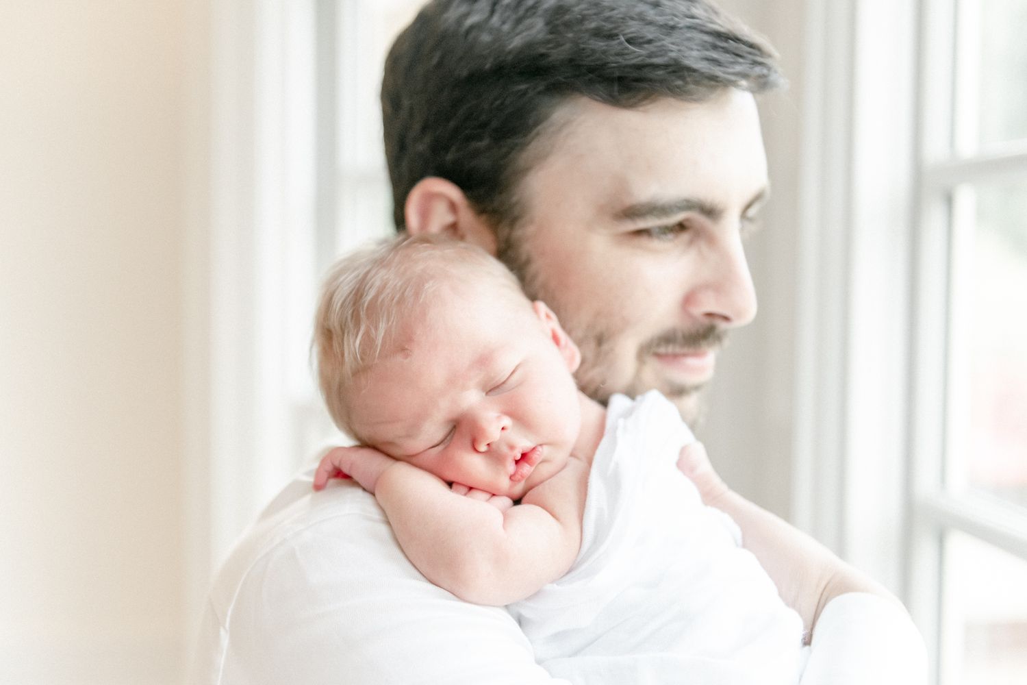 Newborn baby sleeps on his father's shoulder as his father gazes out of the window.