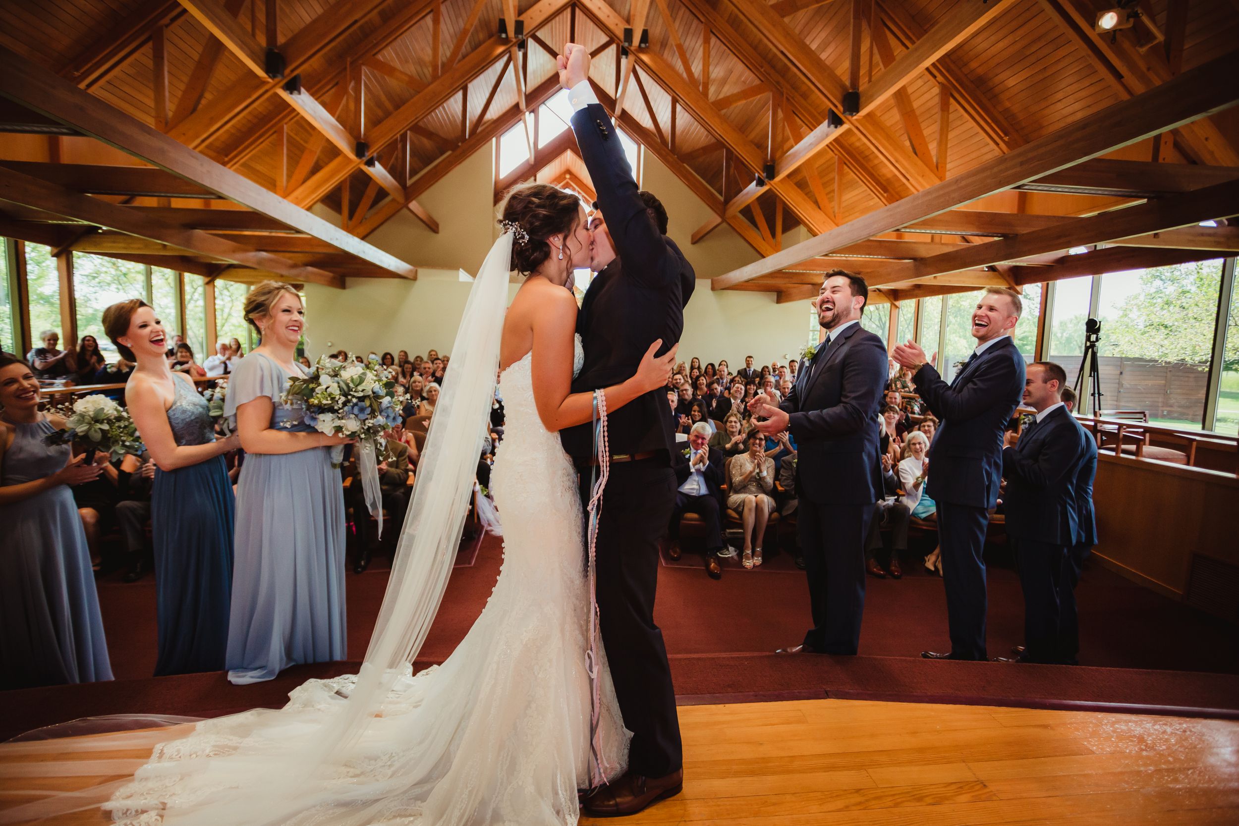 Bride and groom kissing at the end of the ceremony. He has his first in the air. The guests are seen behind them.