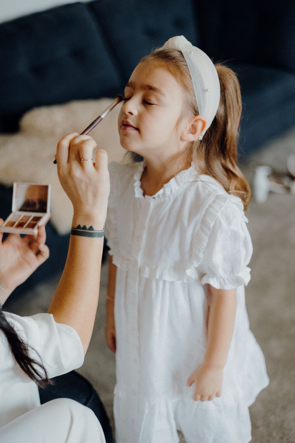 Young girl in white dress getting makeup applied while standing in a living room.