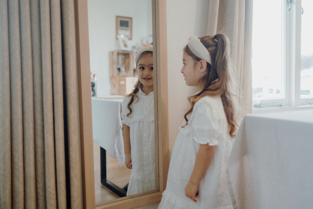 A child in a white dress looks at their reflection in a full-length mirror.