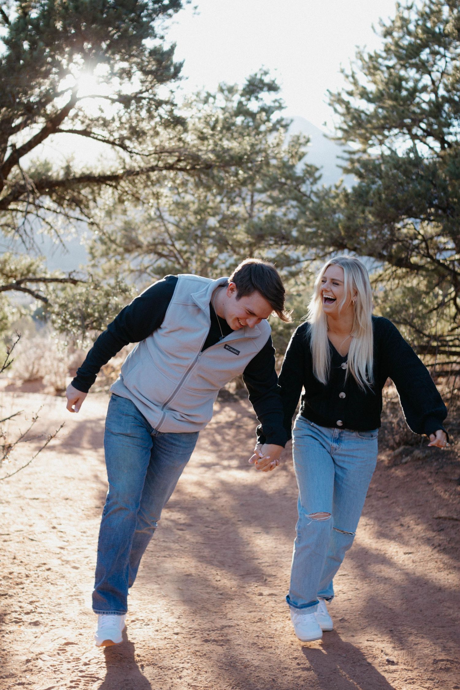 A engaged couple laughing and walking together on a sunny hiking trail with pine trees in the background in Colorado Springs