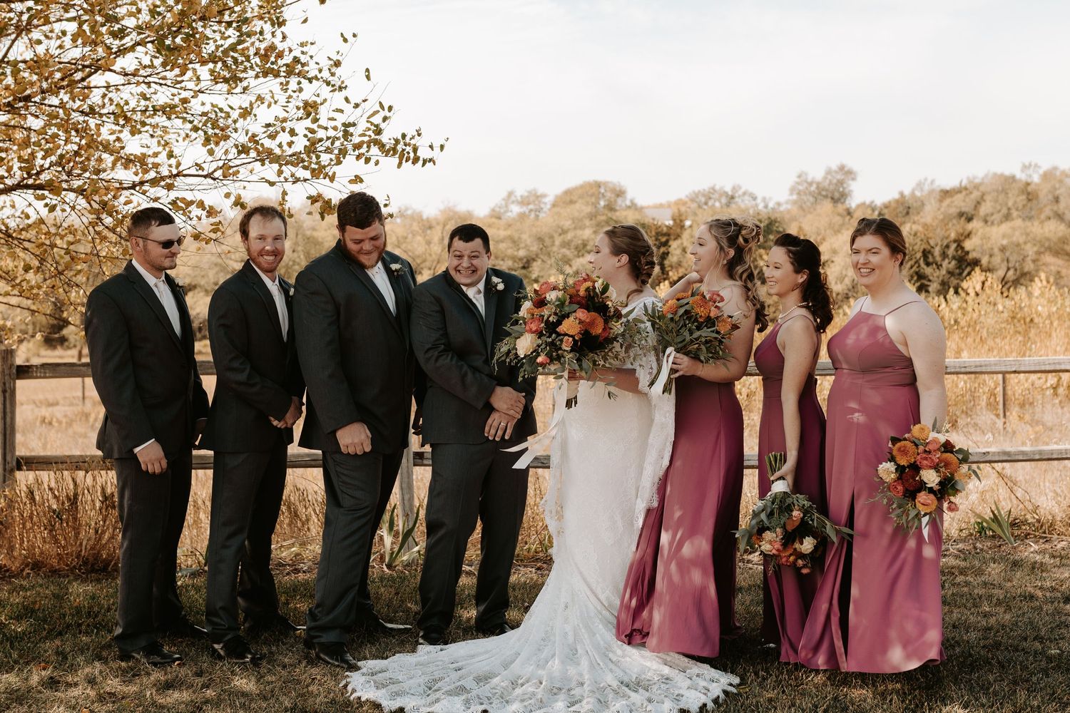 Wedding party in formal attire poses outdoors with bridesmaids in pink dresses and groomsmen in black suits.