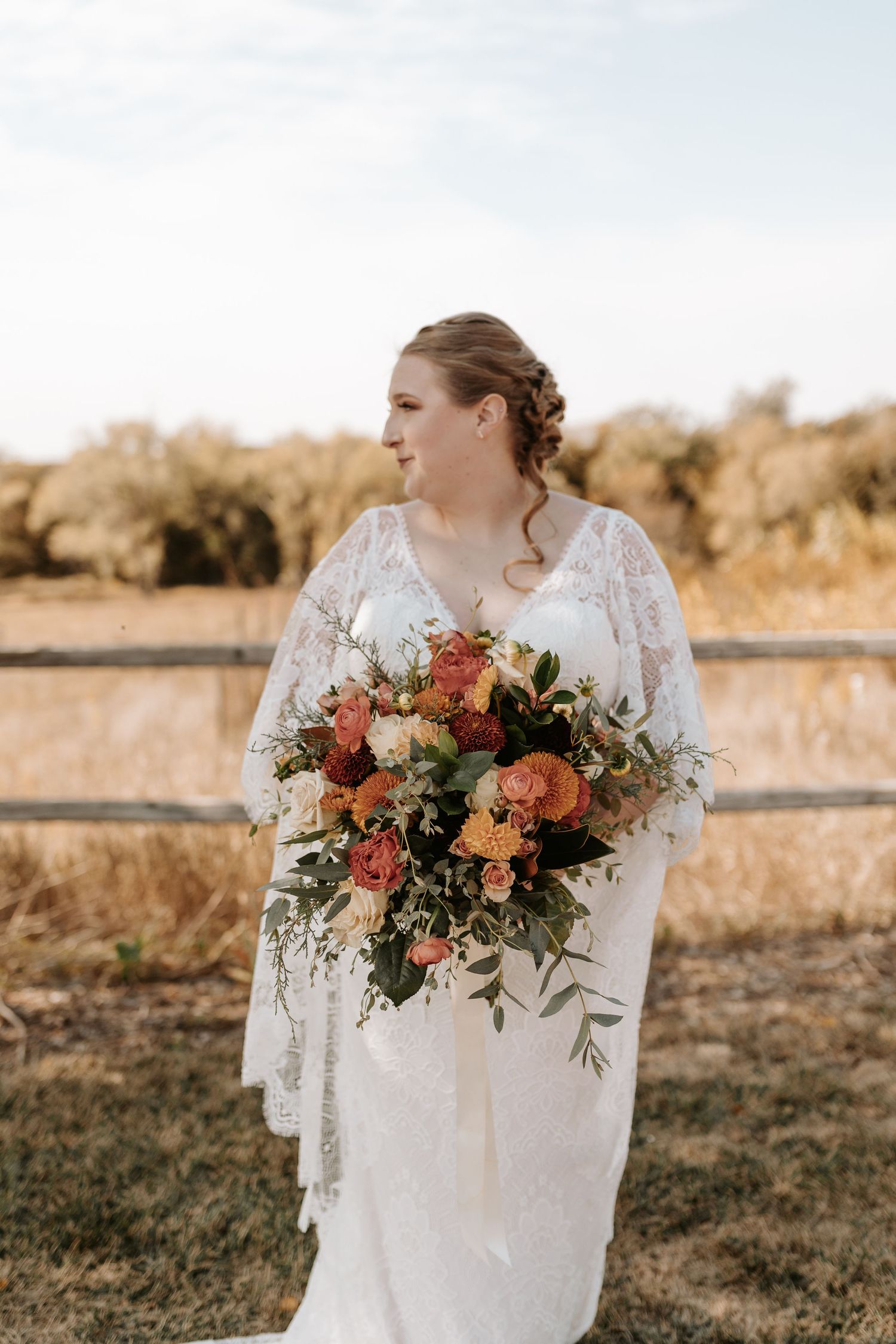 Bride holding vibrant autumn-colored bouquet against rustic farmland backdrop.