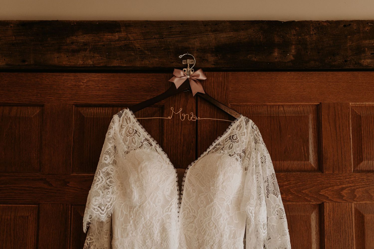 White lace wedding dress with buttons hanging on wooden hanger against dark wood paneling.