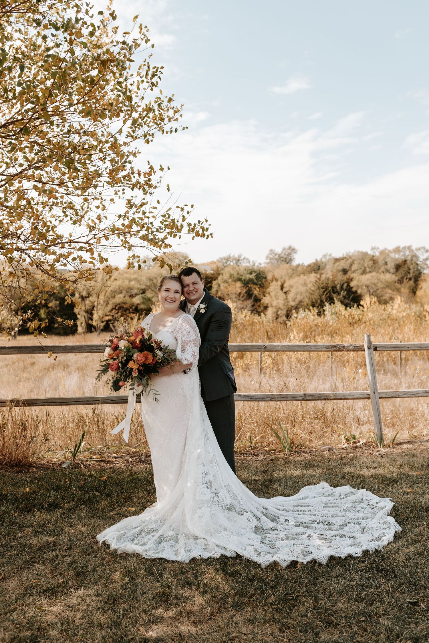 Newlyweds pose against rustic fence with bride's lace train flowing on autumn grass.