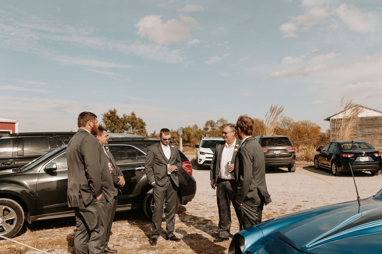 Groomsmen gathered around cars in parking area before wedding ceremony.