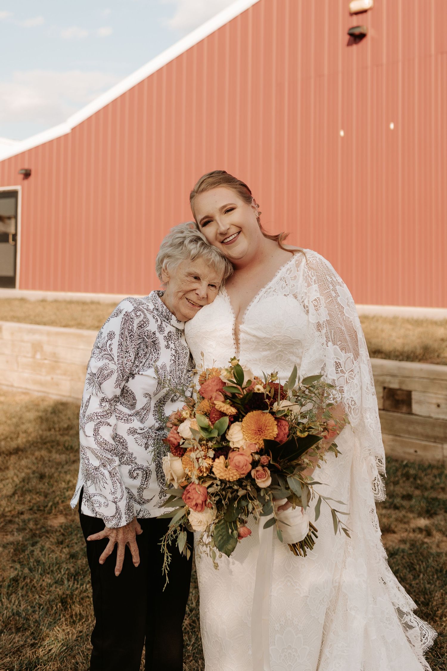 A wedding portrait featuring a traditional lace dress and beautiful peach-toned floral bouquet in front of a rustic barn.