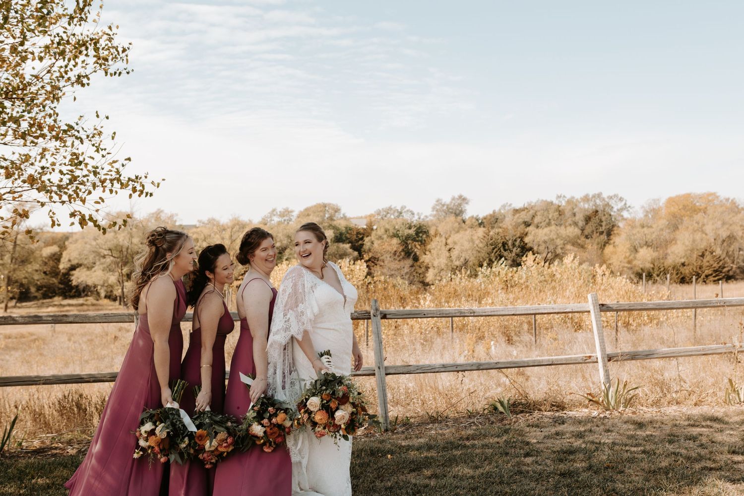 Wedding party in pink dresses posing by rustic fence in golden sunlight.