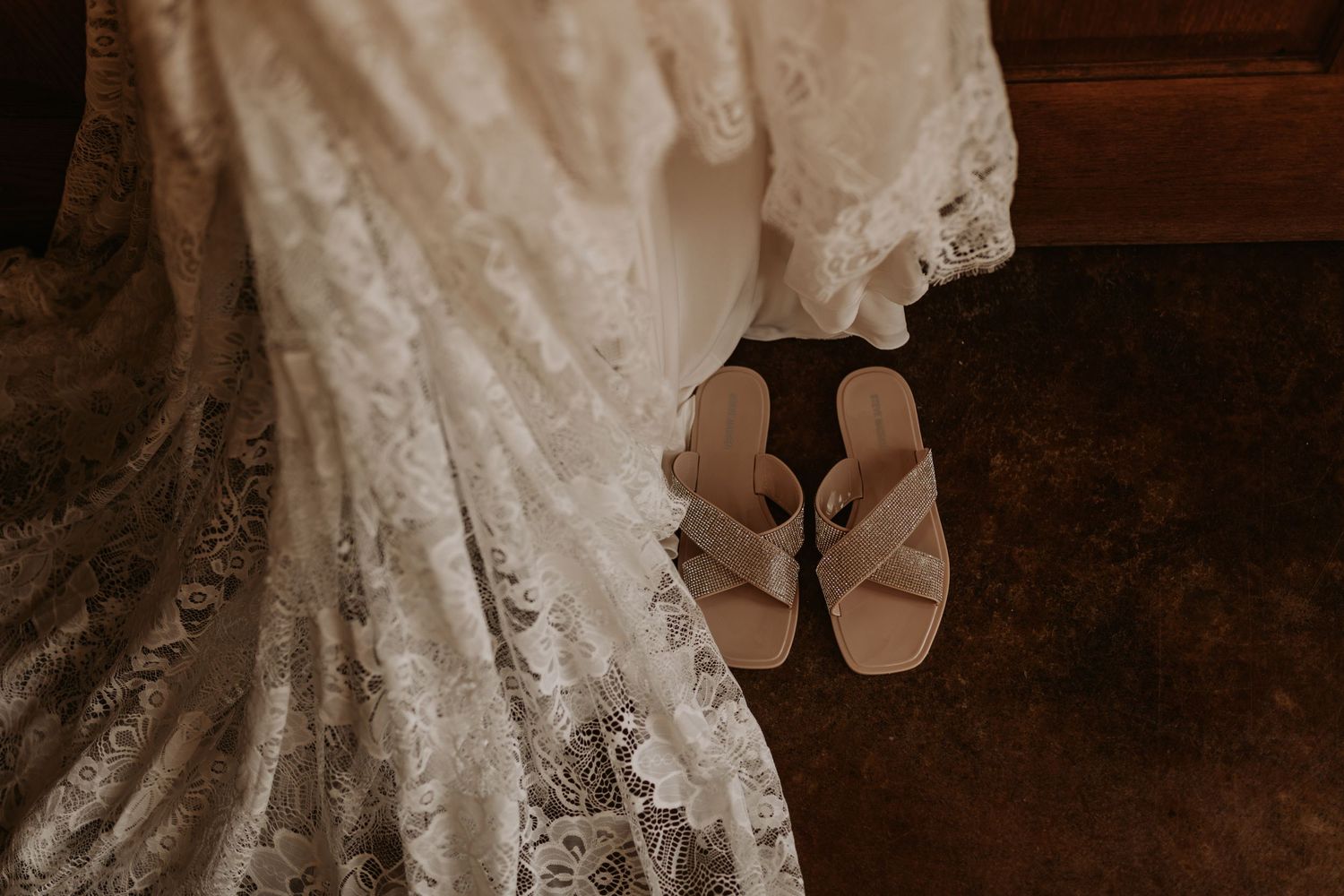Elegant lace wedding dress train with simple sandals on a dark wooden floor.