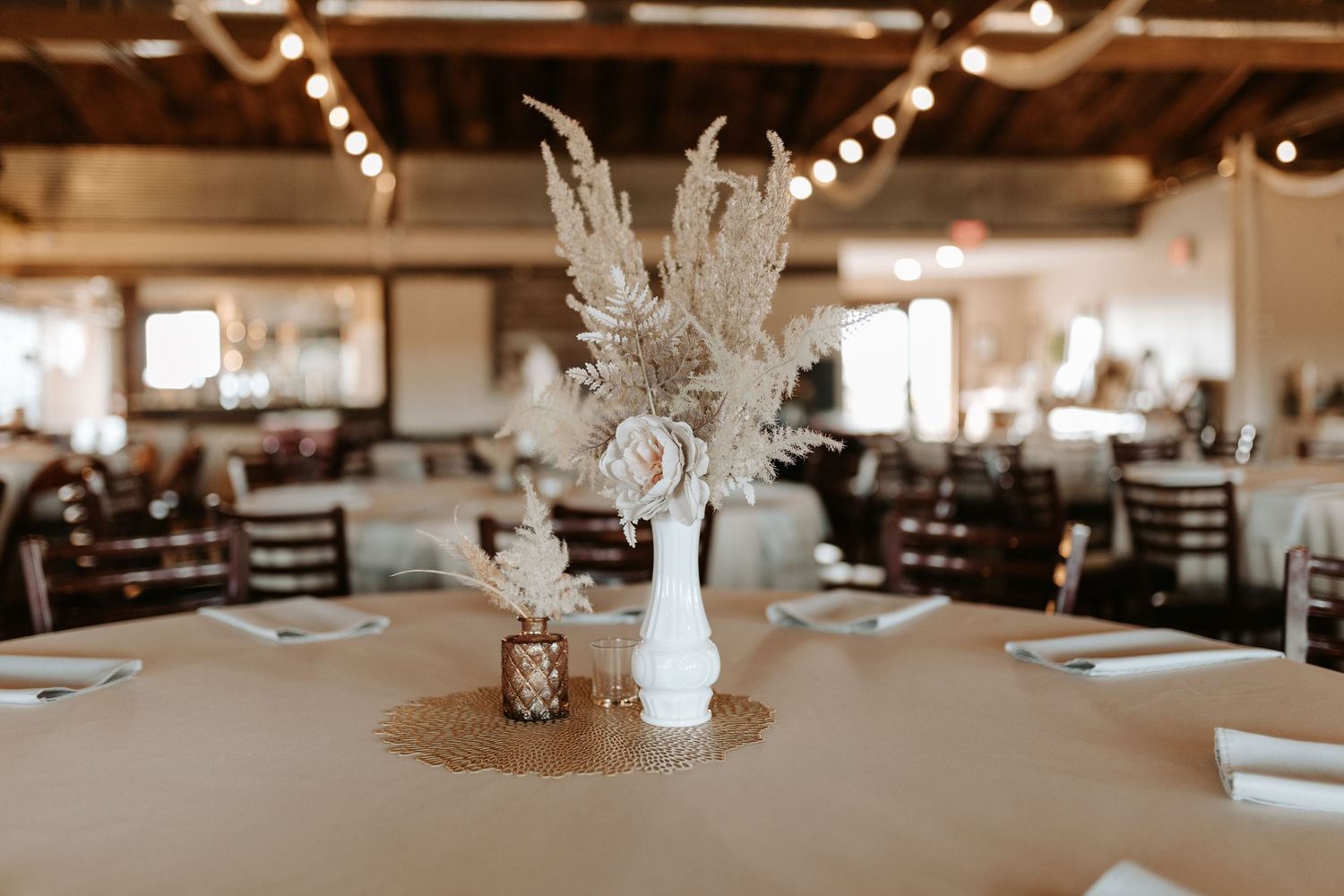 Rustic wedding centerpiece with pampas grass in white vase on table with string lights overhead.