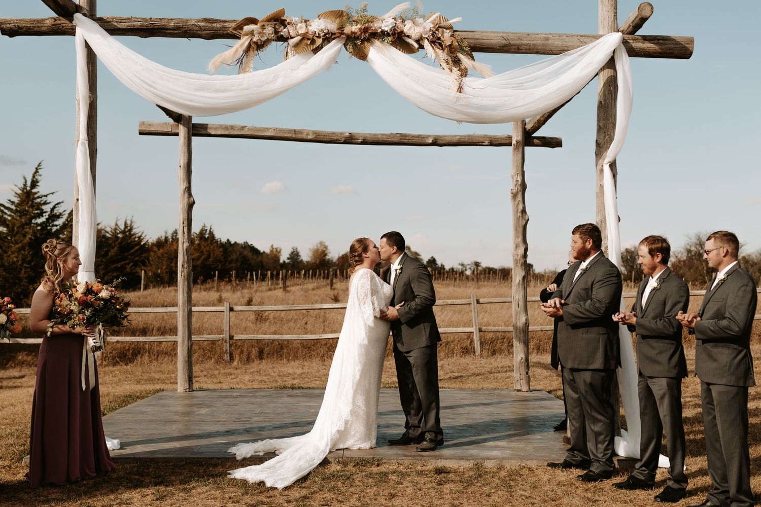 Rustic outdoor wedding ceremony under wooden arch with white fabric and floral decorations.