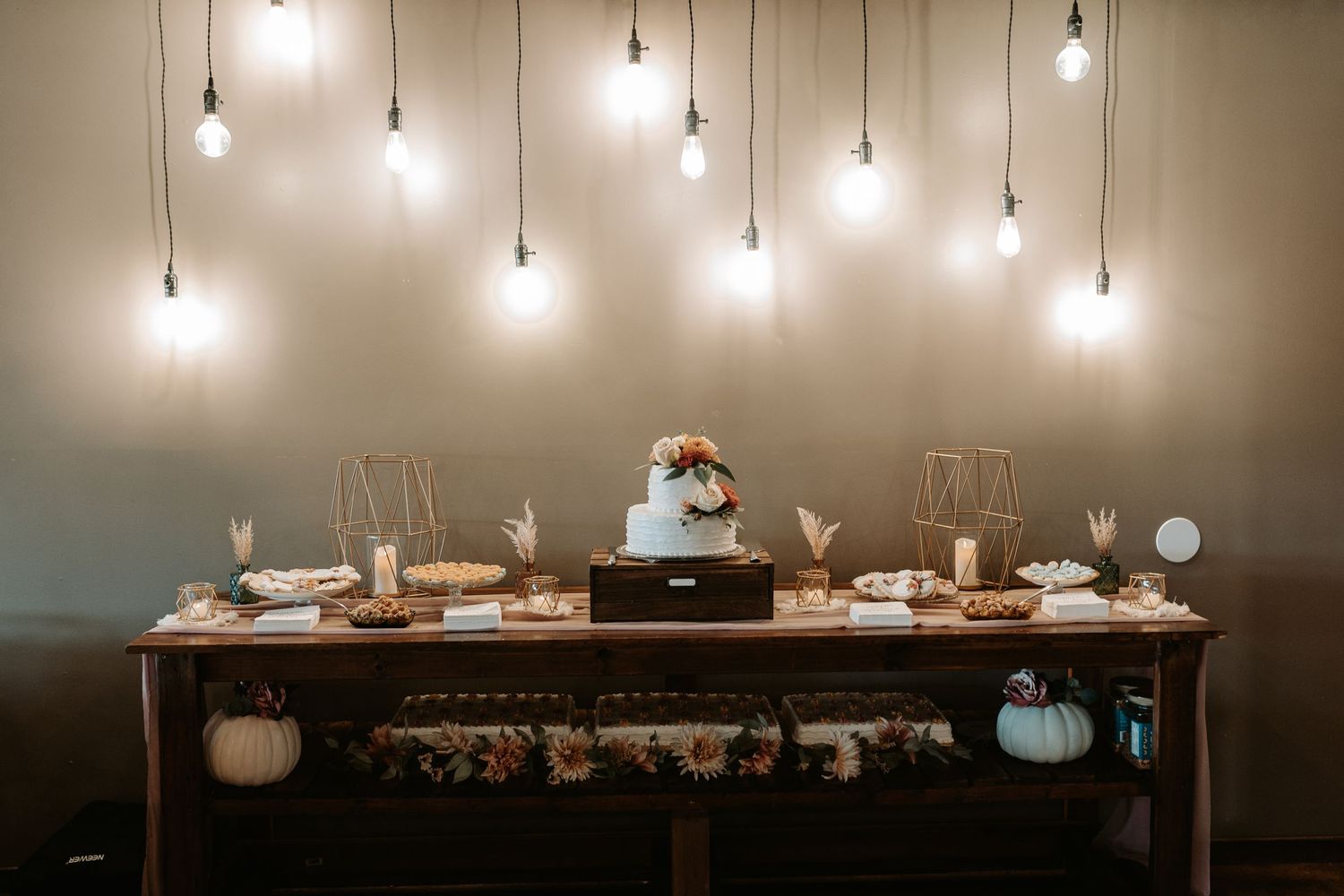 Reception dessert table with hanging lights and white cake surrounded by pumpkin decor.