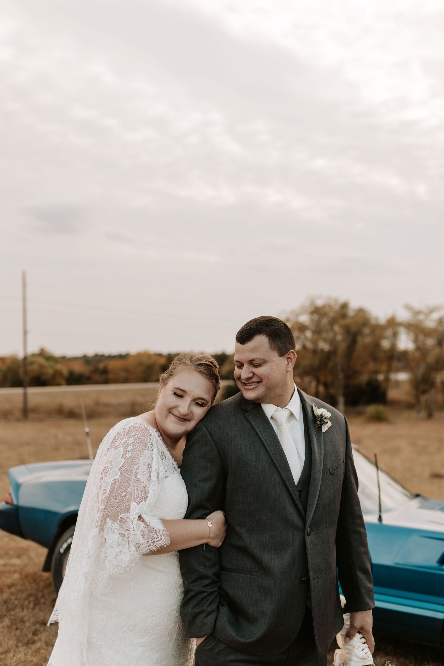 A couple runs hand-in-hand through a golden field with a classic blue car behind them.
