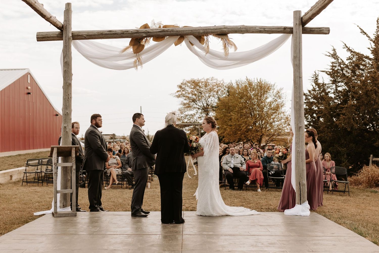 Outdoor wedding ceremony under rustic wooden arch with white fabric draping.