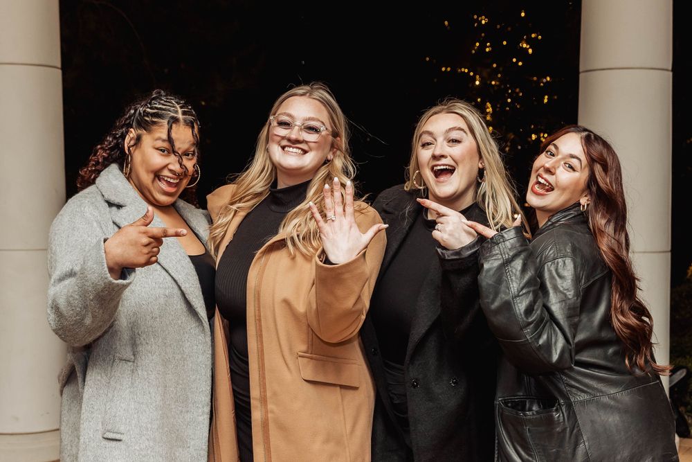 Four friends showing off ring and celebrating together under outdoor lighting.