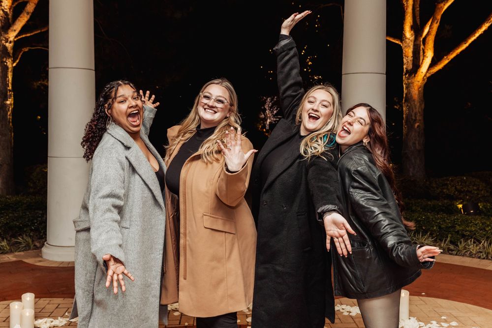 Four friends in winter coats strike dramatic poses while laughing together outdoors at night.