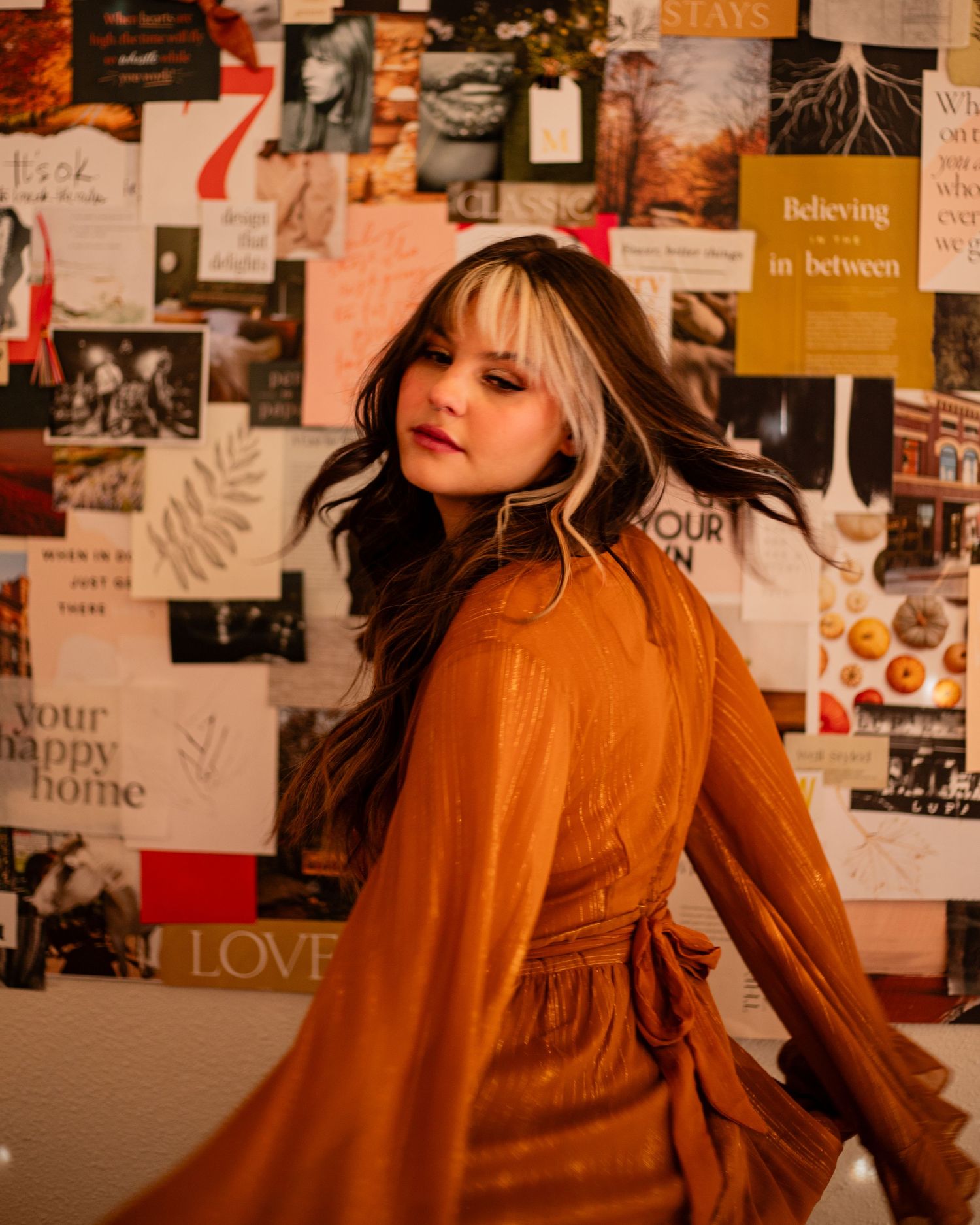 Artistic photoshoot against a wall covered in collages and posters with warm orange tones.
