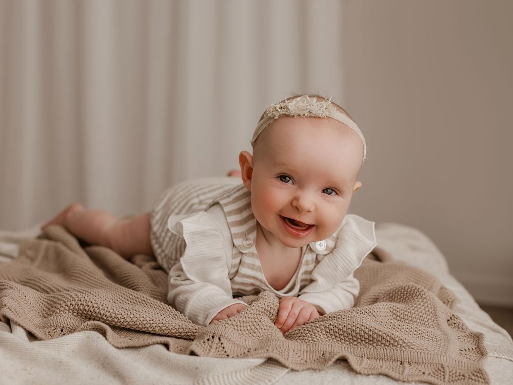 Baby wearing white outfit lies smiling on textured beige blanket during indoor photo session.