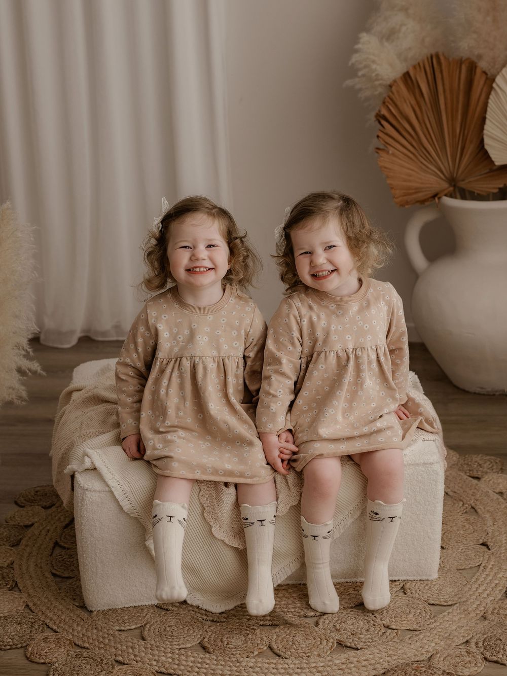 Two matching smiles in identical beige dresses sitting together on cream furniture.