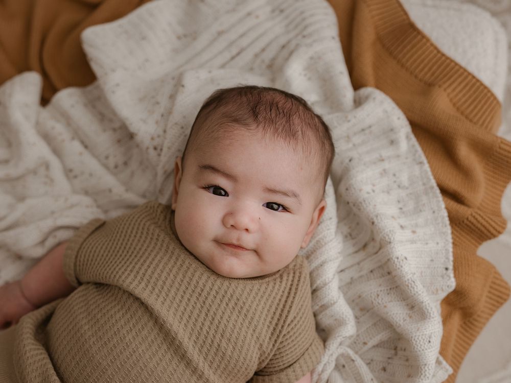 A baby wearing a cozy tan knit sweater lays on a textured white blanket with a tan knit blanket nearby.
