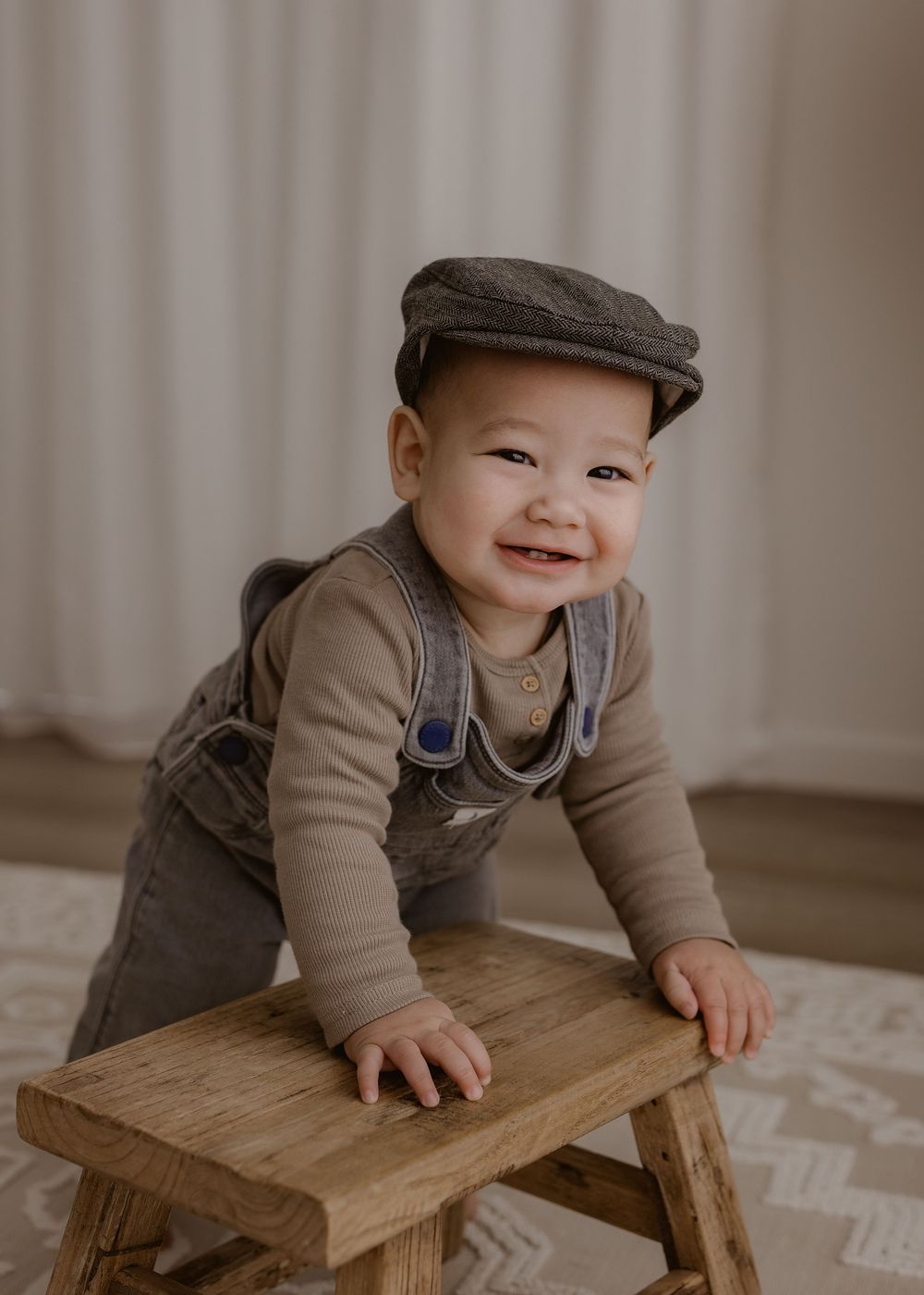A toddler in a brown flat cap and overalls smiles while crawling on a small wooden stool indoors.