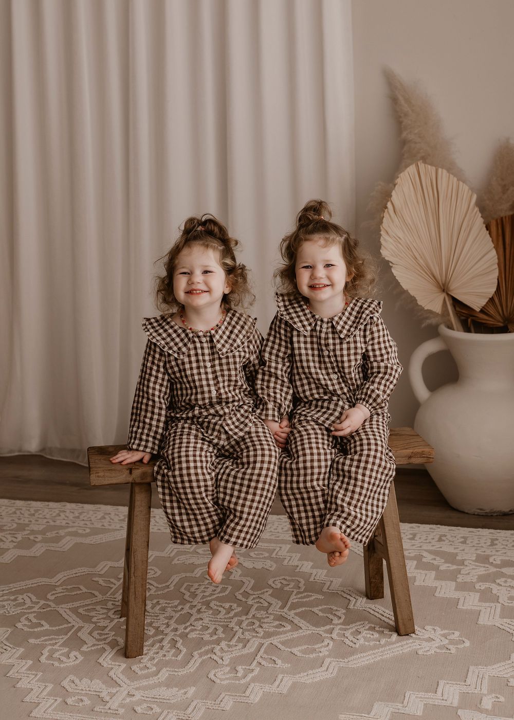 Two children in matching gingham dresses sit and pose playfully on a wooden bench against a neutral background.