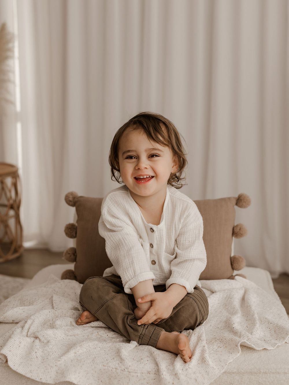 A small child sits cross-legged on a bed wearing a cream colored sweater and brown pants, surrounded by cozy pillows.