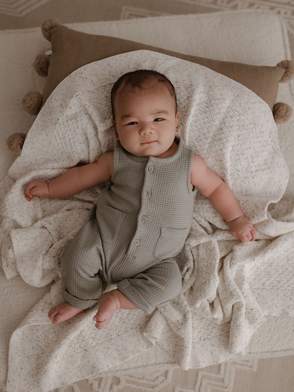 Baby wearing grey textured romper lies on white textured blanket against brown pillow with pom pom trim detail.
