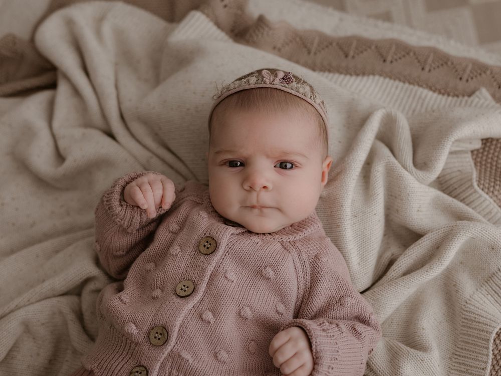 Baby wearing pink knit sweater with wooden buttons and delicate crown lying on beige blanket.