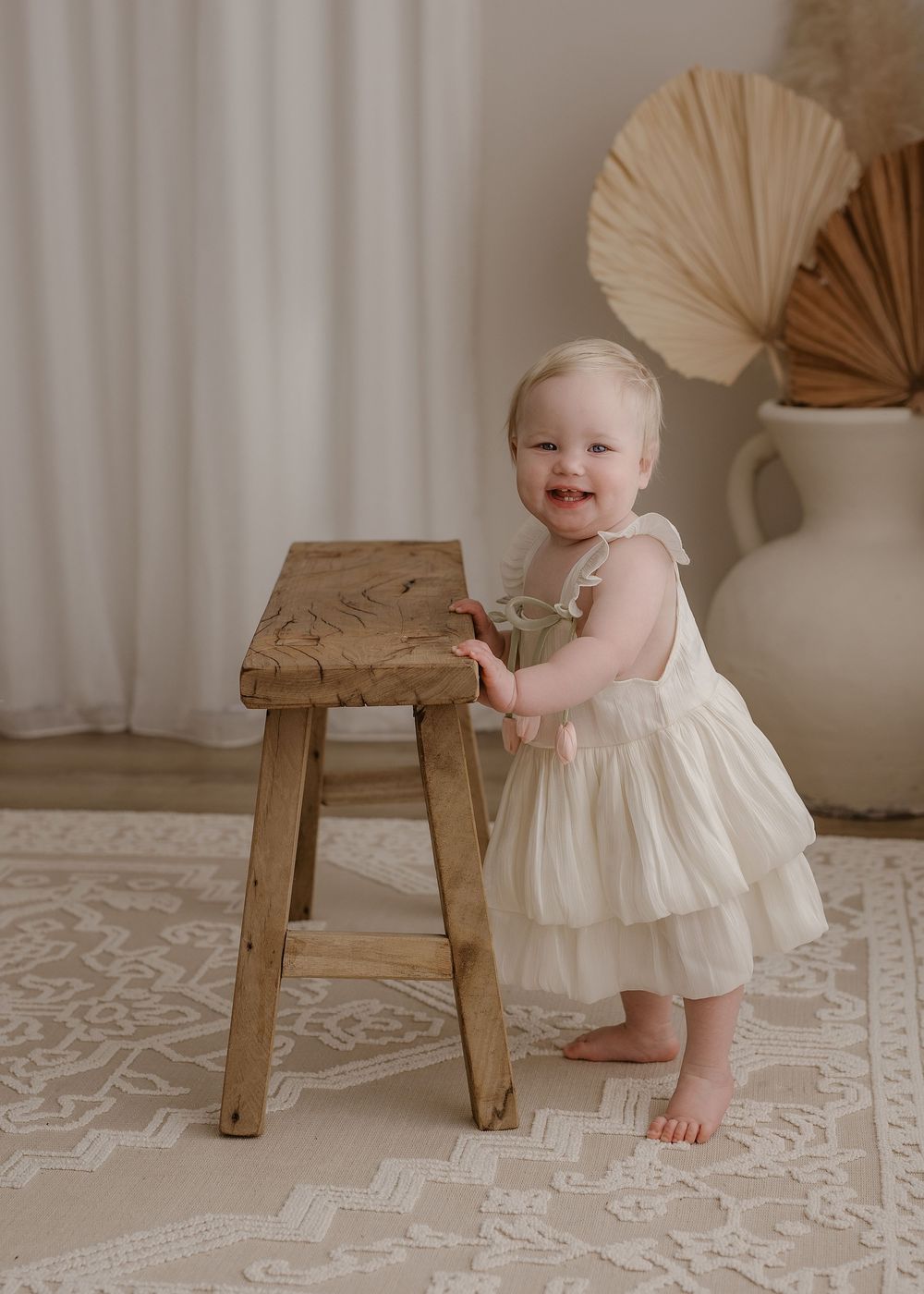 A happy toddler in a white tulle dress stands next to a rustic wooden stool against a neutral backdrop with palm fan decor.