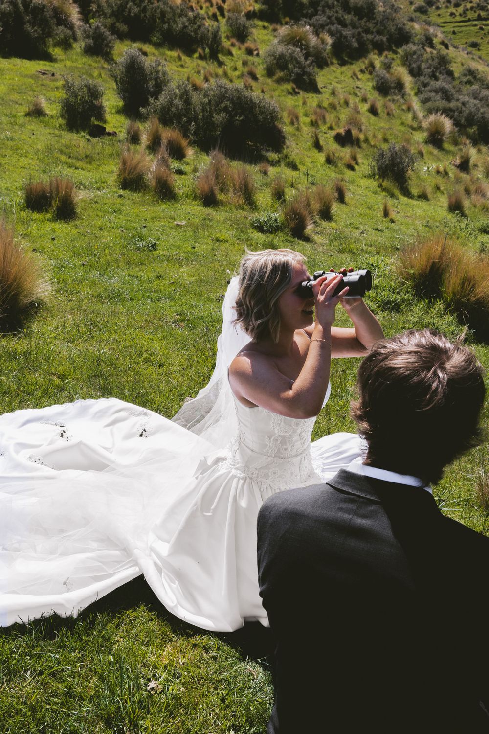 Wedding couple sitting on grassy hillside looking through binoculars together.