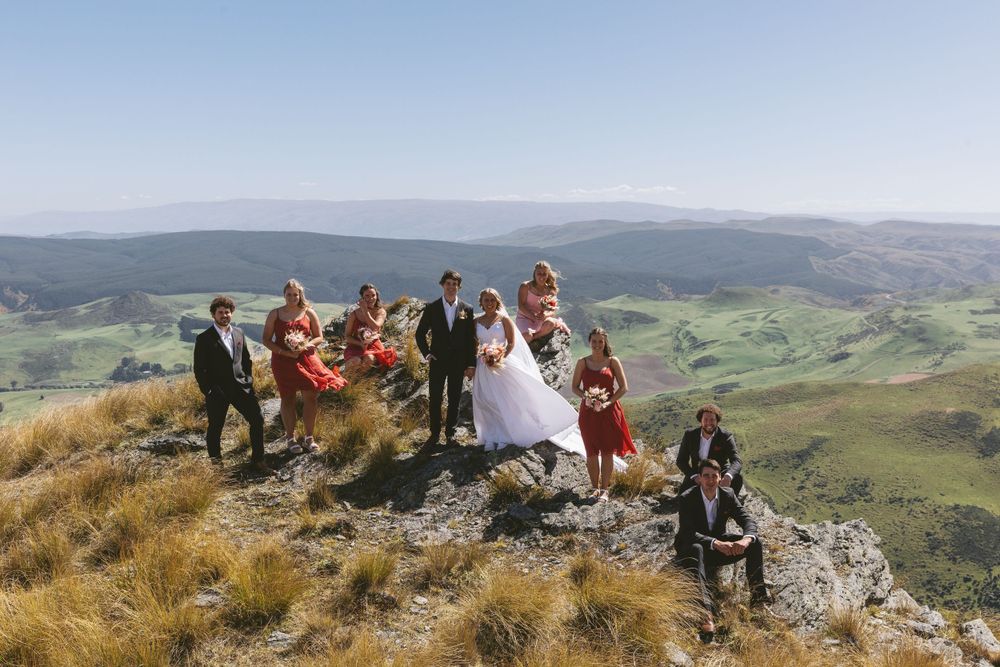 Wedding group poses on rocky outcrop with dramatic valley views and mountains in background.
