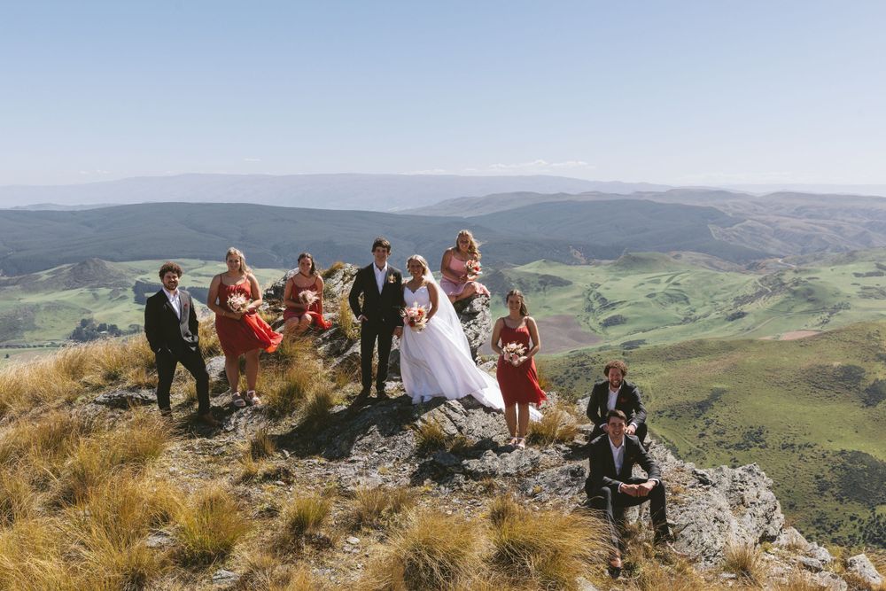Bridal party stands on mountain peak overlooking scenic green valley on clear day.