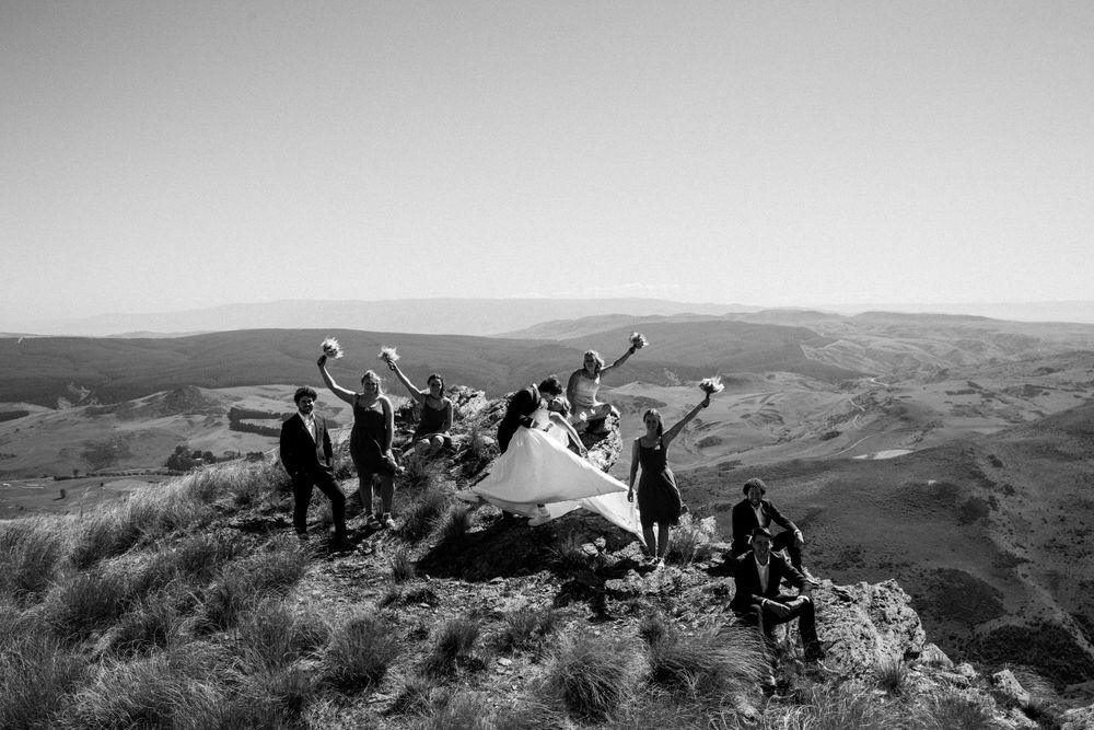Black and white photo of wedding party celebrating on mountain peak with scenic backdrop.